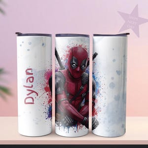 K&ouml;nnte beinhalten: Ein wei&szlig;er Becher mit einem Deadpool-Design. Der Becher hat einen schwarzen Deckel und einen Spritzer roter, blauer und violetter Farbe. Der Name "Dylan" ist vertikal in Rot auf der Seite aufgedruckt.