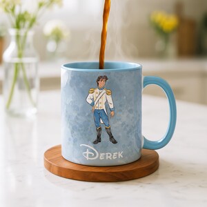 May include: A light blue ceramic mug with a matching handle, featuring a cartoon illustration of a prince in a white and gold uniform. The mug has the name "Derek" printed on it and sits on a wooden coaster.