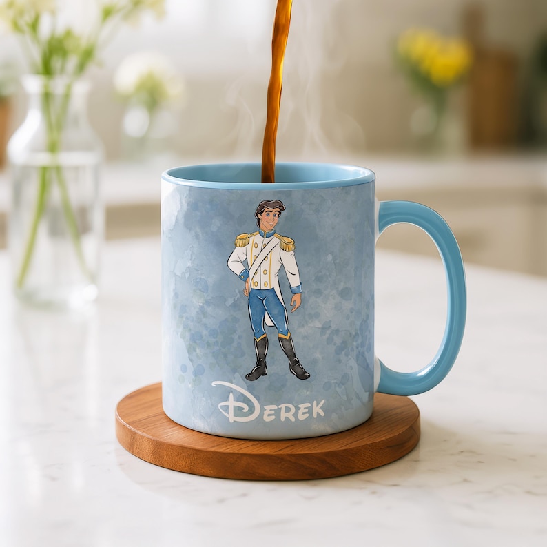 May include: A light blue ceramic mug with a matching handle, featuring a cartoon illustration of a prince in a white and gold uniform. The mug has the name "Derek" printed on it and sits on a wooden coaster.
