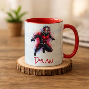 Puede incluir: Taza de cerámica blanca con interior y asa rojos. La taza presenta una colorida ilustración de Ant-Man con el nombre "Dylan" en escritura roja. La taza está sobre un posavasos de madera.