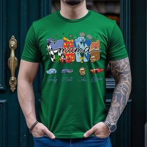Puede incluir: Una camiseta verde con un estampado gr&aacute;fico que incluye la palabra mama en letra cursiva. El dise&ntilde;o presenta letras coloridas decoradas con varios personajes de coches de dibujos animados, banderas a cuadros y los nombres Emily, Matt, John y Olivia.
