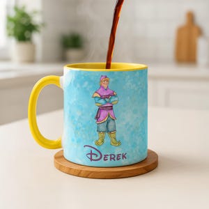 Peut inclure: Une tasse en c&eacute;ramique avec une anse et un int&eacute;rieur jaunes, repr&eacute;sentant une illustration de personnage en tenue violette et bleue. La tasse porte le nom "Derek" dans une police stylis&eacute;e et repose sur un dessous de verre en bois. Du caf&eacute; est vers&eacute; dans la tasse.