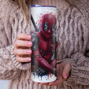 K&ouml;nnte beinhalten: Ein wei&szlig;er Becher mit einer lebendigen Illustration von Deadpool in seinem roten und schwarzen Anzug, mit einem Katana. Das Design beinhaltet Farbspritzer in Rot, Blau und Wei&szlig;. Der Becher wird von einer Person gehalten.