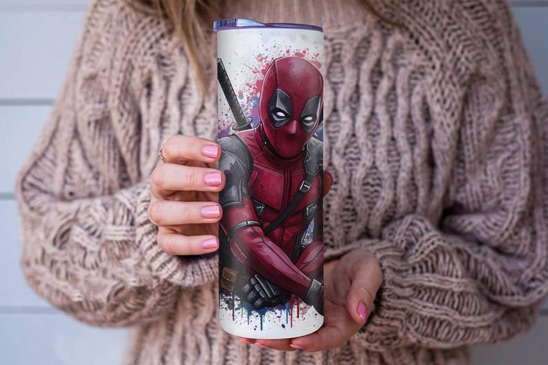 K&ouml;nnte beinhalten: Ein wei&szlig;er Becher mit einer lebendigen Illustration von Deadpool in seinem roten und schwarzen Anzug, mit einem Katana. Das Design beinhaltet Farbspritzer in Rot, Blau und Wei&szlig;. Der Becher wird von einer Person gehalten.
