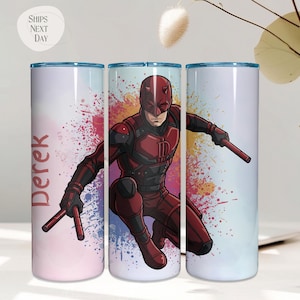 May include: A set of three stainless steel tumblers featuring a superhero illustration. The central tumbler displays a red and black costumed character with batons, set against a colorful paint splatter background. The name "Derek" is printed on one tumbler.