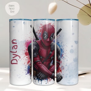K&ouml;nnte beinhalten: Ein wei&szlig;er Becher mit einem Deadpool-Design in einem rot-schwarzen Kost&uuml;m und zwei Schwertern. Der Becher hat einen blauen Rand und den Namen "Dylan" in Rot. Der Hintergrund weist farbenfrohe Farbspritzer auf.