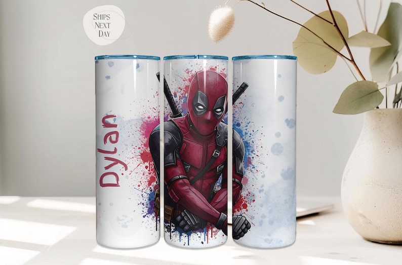 K&ouml;nnte beinhalten: Ein wei&szlig;er Becher mit einem Deadpool-Design in einem rot-schwarzen Kost&uuml;m und zwei Schwertern. Der Becher hat einen blauen Rand und den Namen "Dylan" in Rot. Der Hintergrund weist farbenfrohe Farbspritzer auf.