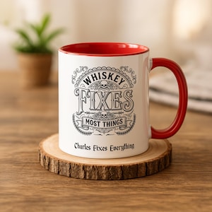 May include: A white ceramic mug with a red interior and handle. The front features black text that reads "WHISKEY FIXES MOST THINGS" with skull and rose illustrations, and the text "Charles Fixes Everything" printed below the main design.