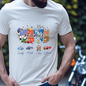 Puede incluir: Una camiseta blanca de manga corta con un estampado gr&aacute;fico que incluye la palabra mama en letra cursiva negra. El dise&ntilde;o presenta personajes de coches de dibujos animados coloridos, banderas a cuadros y estrellas. Debajo, los nombres Emily, Matt, John y Olivia est&aacute;n impresos en azul.