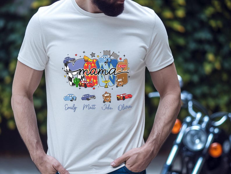 Puede incluir: Una camiseta blanca de manga corta con un estampado gr&aacute;fico que incluye la palabra mama en letra cursiva negra. El dise&ntilde;o presenta personajes de coches de dibujos animados coloridos, banderas a cuadros y estrellas. Debajo, los nombres Emily, Matt, John y Olivia est&aacute;n impresos en azul.
