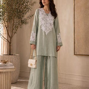 May include: A sage green two-piece outfit featuring a long-sleeved tunic with white lace detailing and wide-leg pants. A small, quilted white handbag with a gold chain strap completes the ensemble. The outfit is suitable for formal or semi-formal occasions.