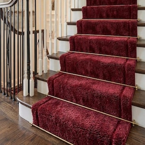 Puede incluir: Una escalera con peldaños de madera oscura y contrahuellas blancas, adornada con una alfombra de felpa de color rojo intenso. La alfombra está asegurada a cada escalón mediante una barra de latón. Una barandilla blanca con pasamanos de madera oscura.