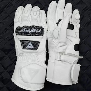 Puede incluir: Un par de guantes de moto blancos con detalles en negro y gris. Los guantes tienen refuerzos en los nudillos, una correa para la muñeca y la marca "DAINESE" es visible. Los guantes son de cuero y tienen un diseño deportivo.