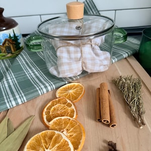 Pode incluir: Uma panela de vidro transparente com alças verdes e um botão de madeira na tampa. Dentro, um feixe embrulhado em tecido xadrez bege e branco. Fatias de laranja seca, paus de canela e raminhos de alecrim são dispostos em uma tábua de corte de madeira.