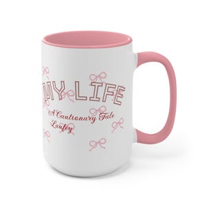 May include: White ceramic mug with a pink interior and handle. The mug features the text "MY LIFE" in a pink, outlined font, along with the phrase "A Cautionary Tale Laufey" and pink bow illustrations.