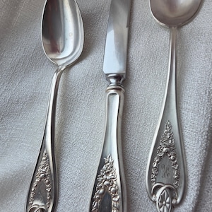 Peut inclure: Trois couverts anciens, dont deux cuillères et un couteau, sont présentés. Les couverts présentent des détails floraux ornés sur les manches et une surface brillante et réfléchissante. Les cuillères sont de forme ovale.