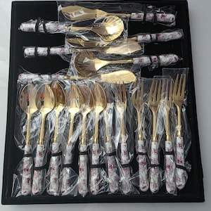 May include: A collection of gold-toned cutlery, comprising spoons and forks, featuring white handles decorated with a floral design. The utensils are presented in a black tray, each piece individually wrapped in clear plastic.