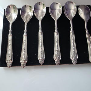 Peut inclure: Ensemble de six cuillères en argent, avec des manches ornés et détaillés. Les cuillères ont un design classique avec un bol arrondi et un motif décoratif sur le manche. Elles sont disposées sur une surface noire.