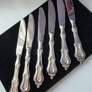 May include: Six silver butter knives with ornate handles are displayed on a black surface. The knives have a shiny, reflective blade and a detailed handle design with gold accents. The handles have a decorative, vintage style.