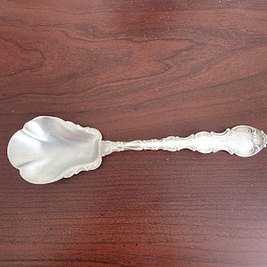 Op de afbeelding: Een zilveren serveerlepel met een sierlijk handvat en een geschulpte kom. De lepel is gemaakt van een glanzend metaal en heeft een gedetailleerd ontwerp op het handvat. De lepel is waarschijnlijk vintage of antiek.