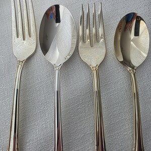 May include: Four pieces of silverware, comprising two forks and two spoons, are arranged on a light-coloured surface. The silverware has a shiny, silver finish with ornate detailing on the handles. The spoons have oval bowls.