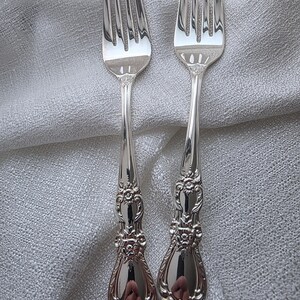 May include: Two silver forks with decorative handles. Each fork has four tines and an ornate floral design on the handle. The forks are presented on a light grey, textured fabric.