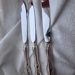 May include: Three silver butter knives with decorative, floral handles. The blades are smooth and reflective, and the handles feature an embossed design. The knives are arranged on a white, textured cloth.