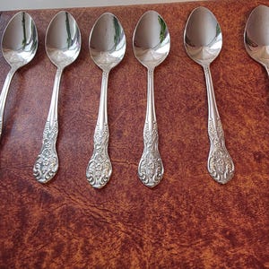 May include: Six silver spoons with ornate floral detailing on the handles are arranged in a row. The spoons have a reflective surface and are set against a brown background. The spoons are likely for serving or eating.