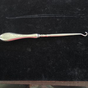 May include: A silver crochet hook with an engraved handle and a curved hook end. The handle features a floral design. This vintage tool is likely for crafting or needlework. The hook is made of metal.