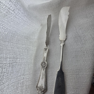 Puede incluir: Dos cuchillos de mantequilla antiguos de plata. Uno tiene un mango decorativo con un diseño ornamentado y las iniciales "S" grabadas. El otro tiene una hoja redondeada y las iniciales "M.G." grabadas en el mango.