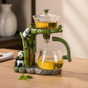 Puede incluir: Infusor de té decorativo con diseño de bambú. La estructura de bambú verde sostiene una tetera y una taza de vidrio. Dos figuras de oso panda están unidas al bambú. El té es de color amarillo dorado.