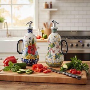 May include: Two decorative ceramic oil bottles with floral designs. One bottle features a red flower with blue and yellow accents, the other has pink and white flowers. Both have black handles and silver spouts. Vegetables and a knife are on a wooden cutting board.