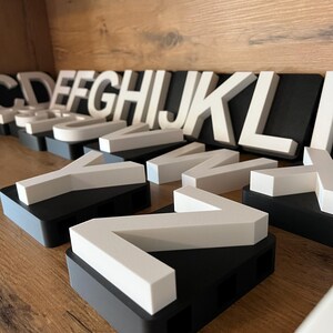 May include: White uppercase letters of the alphabet, mounted on black and gray rectangular bases. The letters are arranged on a wooden shelf. The letters are in a modern, minimalist style.