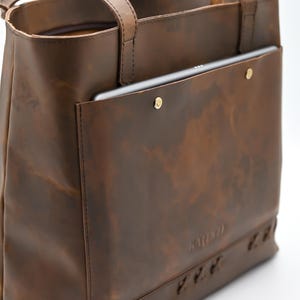 May include: A brown leather tote bag with a tablet in the front pocket. The bag has a handle and a shoulder strap. The leather has a distressed look. The bag is held together with gold rivets.