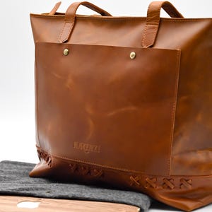 May include: A brown leather tote bag with a large front pocket secured by two brass rivets. The bag has two top handles and a zipper closure. The bottom of the bag features decorative stitching. A gray laptop sleeve is visible below the bag.