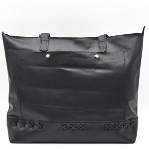 Op de afbeelding: Zwarte leren tote bag met twee handvatten aan de bovenkant en een voorvakje dat is vastgezet met twee goudkleurige klinknagels. De onderkant van de tas heeft decoratieve kruissteekdetails. De tas staat tegen een witte achtergrond.