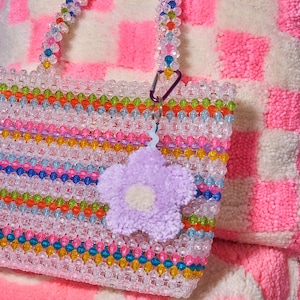 Peut inclure: Un sac fourre-tout en perles multicolores avec un charme en forme de fleur violette. Le sac a une base transparente et est fait de petites perles colorées. Le charme de la fleur est attaché à un clip argenté.