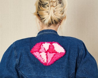 May include: A blue denim jacket with a pink and white yarn diamond shape on the back.