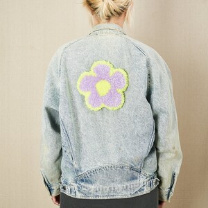 Puede incluir: Una chaqueta vaquera azul claro con un gran parche de flor morado y amarillo en la espalda.