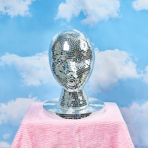 May include: A silver disco ball head sculpture on a pink fuzzy surface. The disco ball is made of many small mirrored tiles.