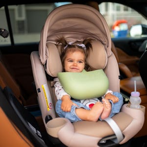 May include: A tan and beige car seat with a baby inside. The baby is wearing a green bib and blue jeans. A sippy cup is in the cup holder. The car seat is installed in a car.