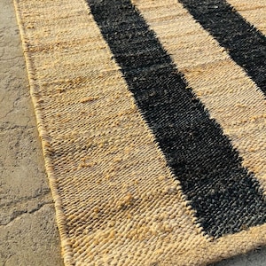 K&ouml;nnte beinhalten: Ein rechteckiger Webteppich mit einem wiederkehrenden Muster aus breiten, naturfarbenen Jute-Fasern und kr&auml;ftigen schwarzen Streifen. Die Textur ist grob und organisch, mit einer sichtbaren Flechtstruktur &uuml;ber die gesamte Oberfl&auml;che.