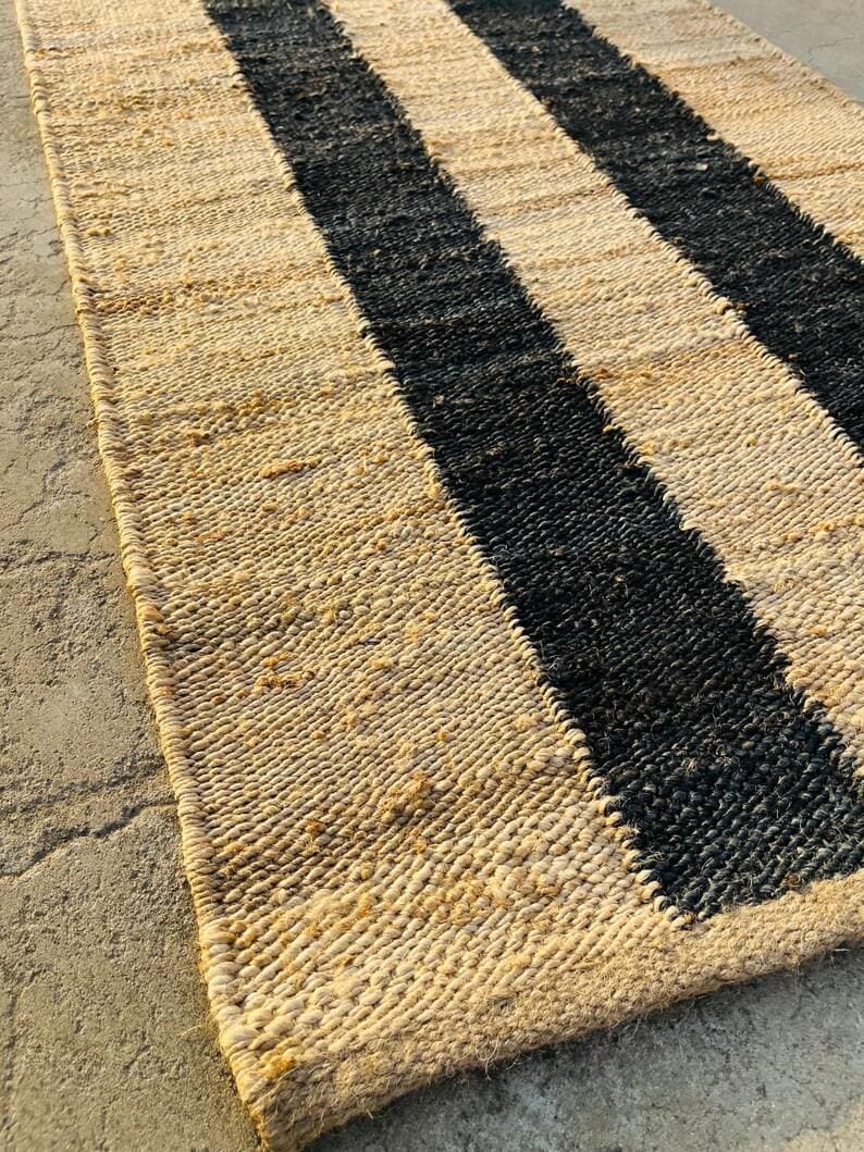 K&ouml;nnte beinhalten: Ein rechteckiger Webteppich mit einem wiederkehrenden Muster aus breiten, naturfarbenen Jute-Fasern und kr&auml;ftigen schwarzen Streifen. Die Textur ist grob und organisch, mit einer sichtbaren Flechtstruktur &uuml;ber die gesamte Oberfl&auml;che.