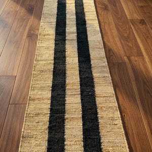 K&ouml;nnte beinhalten: Ein rechteckiger L&auml;ufer aus naturfarbenen, gewebten Fasern mit zwei markanten, parallelen schwarzen Streifen, die &uuml;ber die gesamte L&auml;nge verlaufen. Die Textur wirkt grob und organisch, mit einer flachen und kompakten Konstruktion.