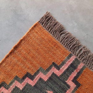 May include: A woven rug featuring a vibrant orange base with a geometric pattern in dark gray and soft pink. The rug has a textured, coarse weave and is finished with a long, frayed fringe along the edge, creating a distinct decorative border.
