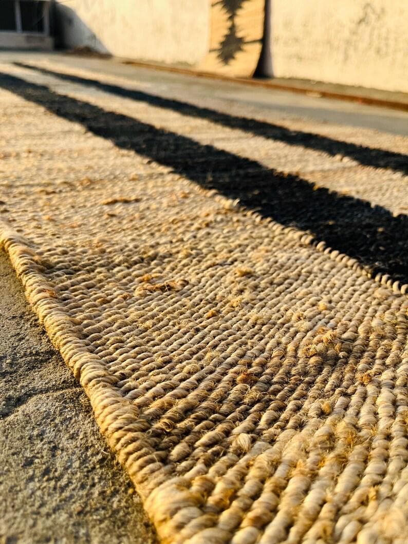 K&ouml;nnte beinhalten: Ein rechteckiger Webteppich mit einer nat&uuml;rlichen beigen Faserstruktur und einem markanten, dicken schwarzen L&auml;ngsstreifen. Der Teppich weist einen geflochtenen Randabschluss sowie eine dichte, grobe Struktur aus nat&uuml;rlichen Pflanzenfasern auf.