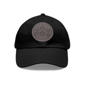 May include: Black baseball cap with a circular patch featuring dice illustrations. The patch is gray with a stitched border and includes various dice shapes and numbers. The cap has a curved brim and a structured crown.