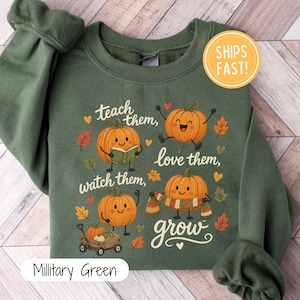 Könnte beinhalten: Ein militärgrünes Langarm-Sweatshirt mit einer Grafik von vier orangefarbenen Kürbissen mit Gesichtern. Die Kürbisse sind beim Lesen eines Buches, beim Springen, Stehen und mit einem Schal dargestellt. Der Text lautet: teach them, love them, watch them, grow.