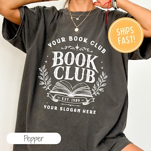 Könnte beinhalten: Ein anthrazitfarbenes Kurzarm-T-Shirt mit einem weißen Grafikdruck, der YOUR BOOK CLUB, BOOK CLUB, EST. 1989 und YOUR SLOGAN HERE zeigt, zentriert um eine Illustration eines offenen Buches, eingerahmt von dekorativen Blättern und Sternen.