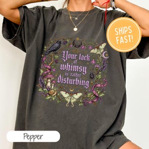 Könnte beinhalten: Ein anthrazitfarbenes Kurzarm-T-Shirt mit einem Grafikdruck und dem Text Your lack of whimsy is rather disturbing. Das Design zeigt einen schwarzen Raben, Motten, Pilze, eine Schnecke, einen Käfer und einen Halbmond umgeben von Ranken.